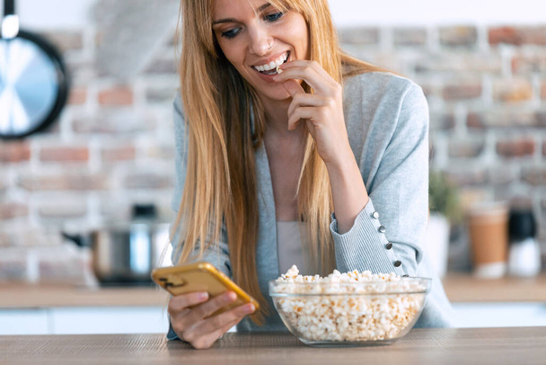 Aufnahme einer lächelnden Frau, die Popcorn aus einer Schüssel greift und pflückt, während sie ihr Mobiltelefon zu Hause benutzt. - Foto, Bild