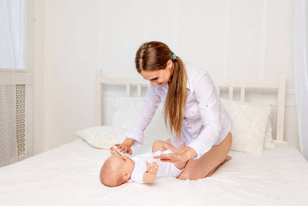 mother sits with baby 6 months on a white bed at home and plays with it, place for text - Foto, Imagen