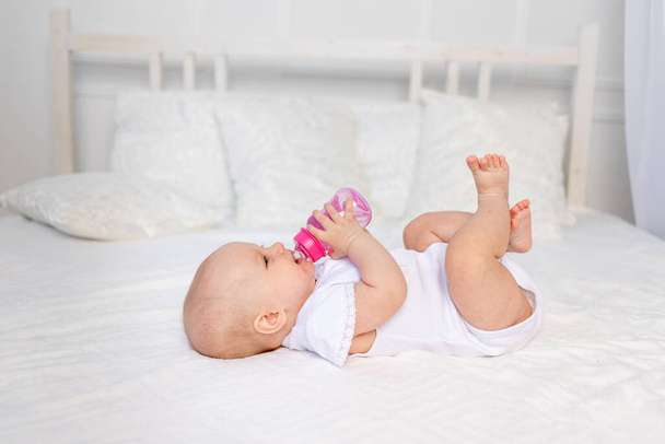 egy 6 hónapos csecsemő fekszik egy fehér ágyon, és iszik tejet egy üvegből, bébiétel koncepció, szöveges hely - Fotó, kép