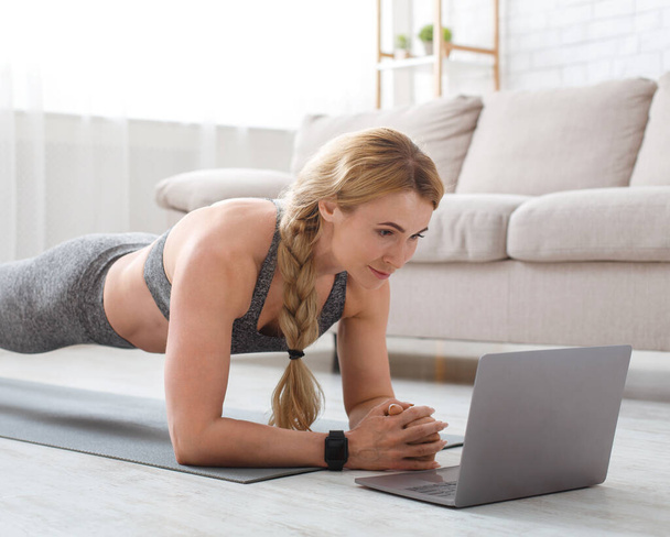 Middle aged woman in sportswear with fitness tracker makes plank at mat on floor and looks at laptop - Zdjęcie, obraz