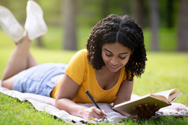 Estudiante negra tomando notas de libro de texto sobre manta de picnic en el parque
 - Foto, imagen