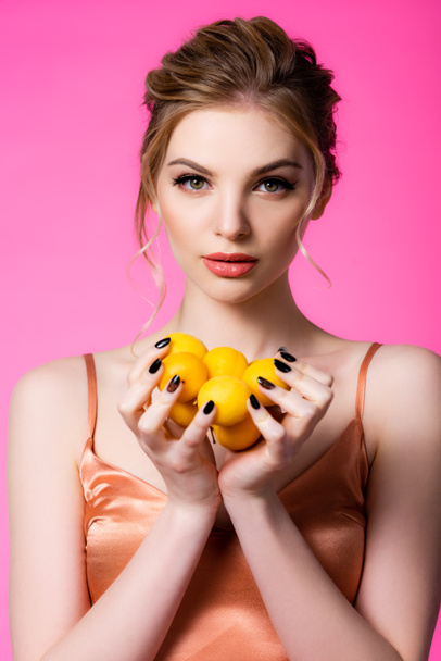 elegant beautiful blonde woman in silk dress holding ripe apricots isolated on pink - 写真・画像