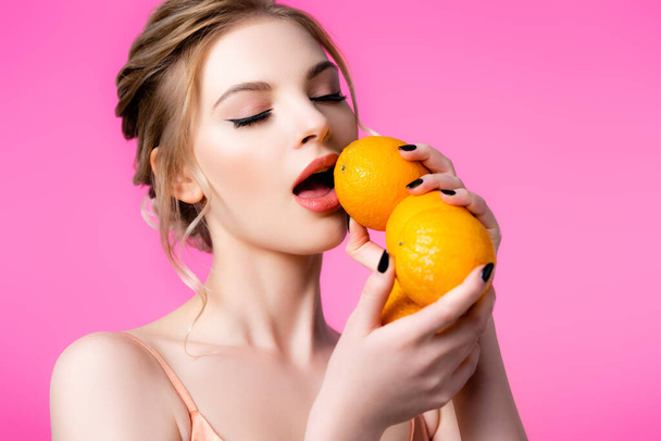 elegant beautiful blonde woman holding ripe oranges near mouth isolated on pink - Photo, image
