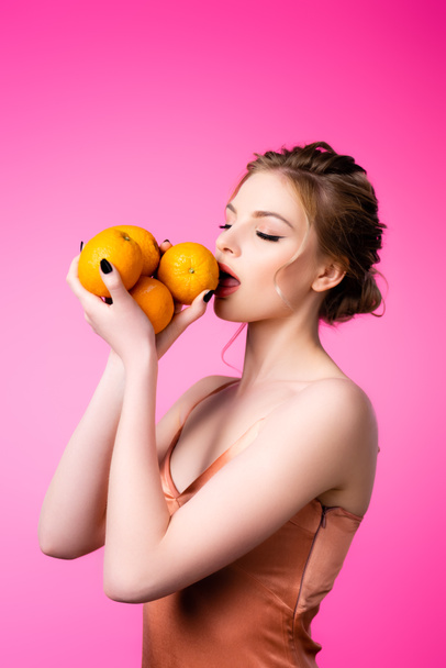 κομψή όμορφη ξανθιά γυναίκα γλείφει ώριμα πορτοκάλια απομονωμένα σε ροζ - Φωτογραφία, εικόνα