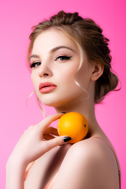 elegant beautiful blonde woman holding ripe orange on shoulder isolated on pink - Fotoğraf, Görsel