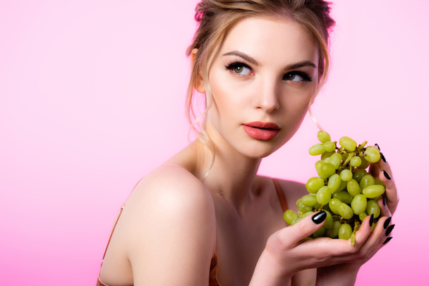 élégante belle femme blonde tenant des raisins verts et regardant loin isolé sur rose - Photo, image