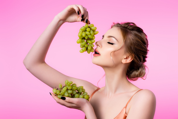 elegant beautiful blonde woman holding green grapes near mouth isolated on pink - Fotografie, Obrázek