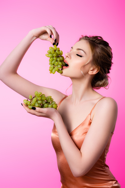 elegant beautiful blonde woman eating green grapes isolated on pink - Fotó, kép