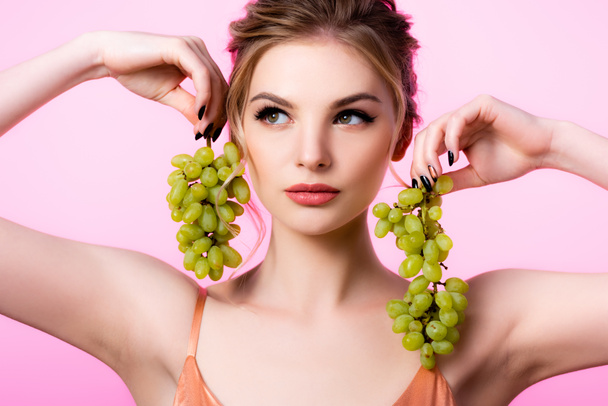 elegante hermosa mujer rubia sosteniendo uvas verdes cerca de la cara aislado en rosa
 - Foto, imagen