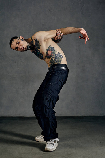 Athletic tattooed male with naked torso, beard. Dressed in black pants and white sneakers. Dancing against gray background. Dancehall, hip-hop - Zdjęcie, obraz