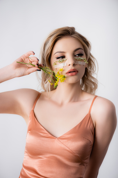 beautiful blonde woman in silk dress with wildflowers under eyes and near mouth isolated on white - Photo, Image
