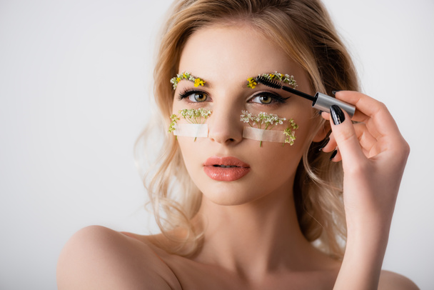 naked beautiful blonde woman styling wildflowers on eyebrows with brow gel isolated on white - Photo, Image