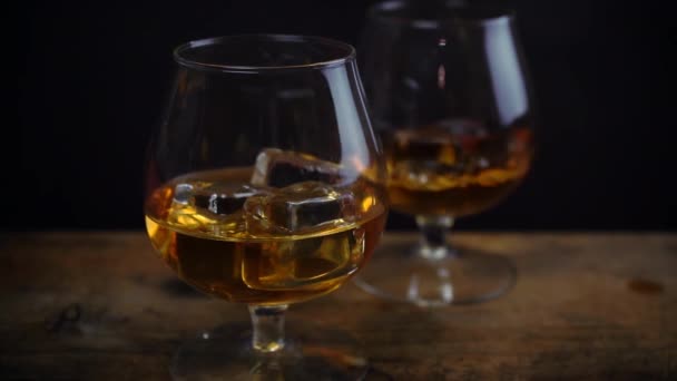 Cubo di ghiaccio che cade nel bicchiere di whisky cognac, colore dorato forte alcol spirito in bicchieri di cognac su sfondo nero scuro, riprese al rallentatore - Filmati, video