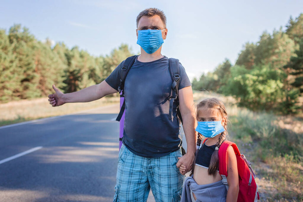 Local traveler. Middle aged father with his daughter with backpacks and wearing medical masks stopping car on the road during their local vacation, new normal summertime, active outdoor lifestyle - Photo, Image