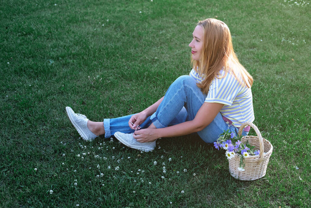 Piękna kobieta siedząca na trawie i szyjąca trampki. szczęśliwa uśmiechnięta kobieta w średnim wieku z koszem kwiatów relaksująca się na trawie. Koncepcja wakacji letnich. - Zdjęcie, obraz