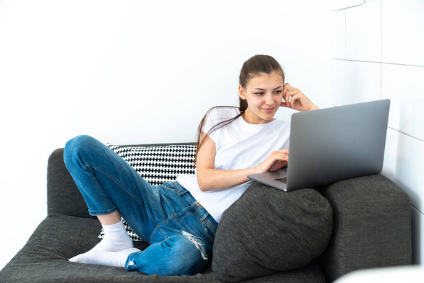Genç, çekici, beyaz tenli esmer, oturma odasında kanepede oturuyor ve bilgisayarını serbest çalışmak için kullanıyor. - Fotoğraf, Görsel