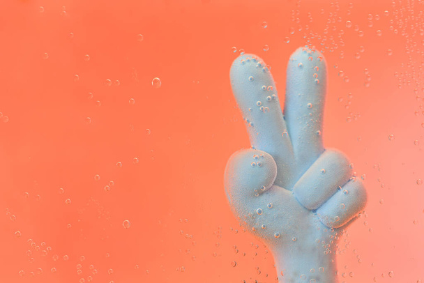Ένα μπλε πλαστικό χέρι κολυμπάει κάτω από το πορτοκαλί νερό και κάνει το σημάδι της νίκης - Φωτογραφία, εικόνα