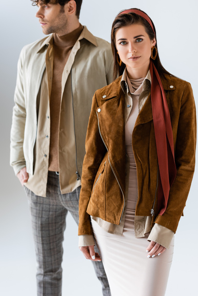 селективный фокус привлекательной женщины, смотрящей в камеру рядом с мужчиной, смотрящей в стильную осеннюю одежду, изолированную на сером - Фото, изображение
