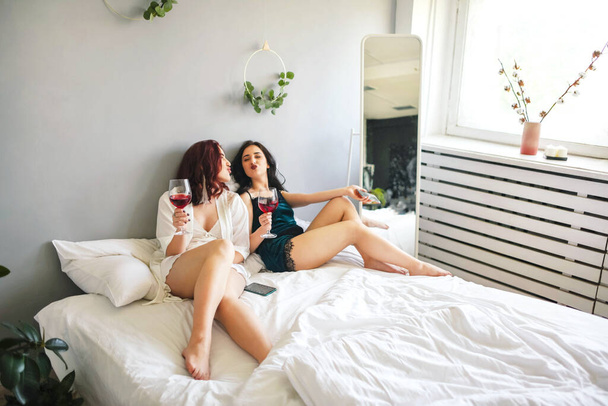 Les copines en pyjama sont assises sur le lit avec du vin et des téléphones.  - Photo, image