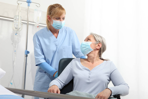 медсестра приймає комфортну літню жінку в інвалідному візку в хірургічних захисних медичних масках в лікарняній кімнаті, концепція ізоляції від коронного вірусу 19
 - Фото, зображення