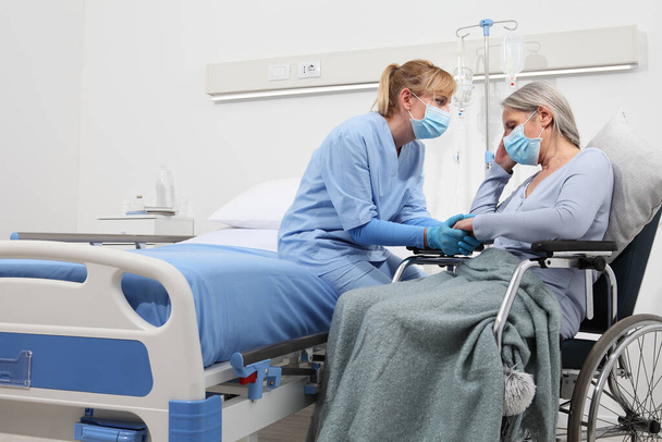 nővér vigaszt nyújt idős nő tolószékben visel sebészeti védő orvosi maszkok kórházi szobában, koncepció izoláció koronavírus covid 19 - Fotó, kép