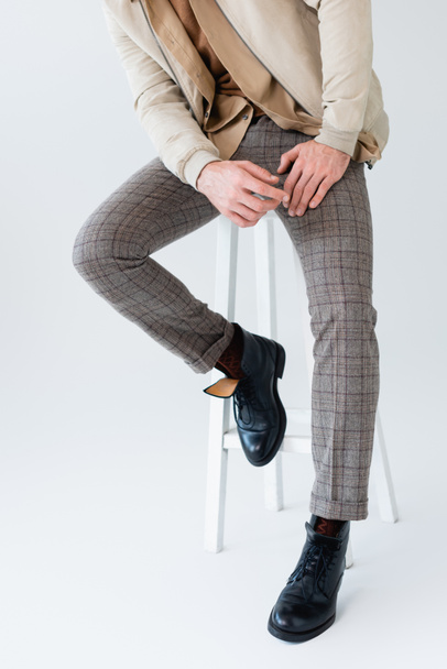 cropped view of trendy man in grey trousers and black boots sitting on stool on grey - Фото, зображення
