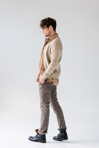 side view of trendy man in autumn clothes posing with hands in pockets on grey - Foto, Imagen