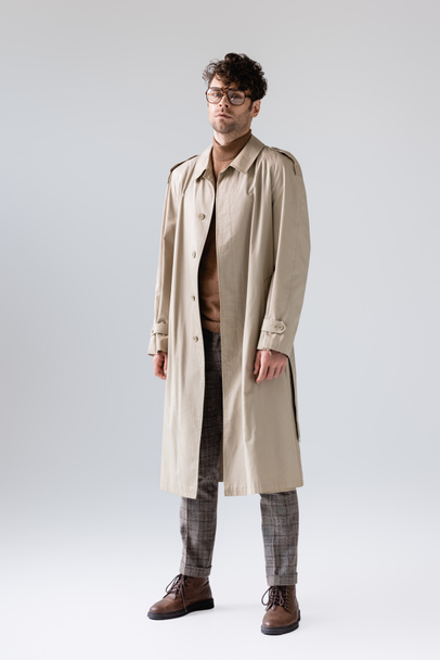 visão de comprimento total do homem na moda em casaco de trincheira olhando para a câmera enquanto posando em cinza
 - Foto, Imagem