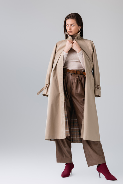 full length view of fashionable girl touching trench coat and looking away on grey - Foto, Imagen