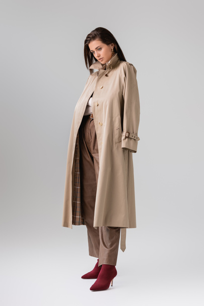 full length view of stylish girl with trench coat on shoulders posing on grey - Zdjęcie, obraz
