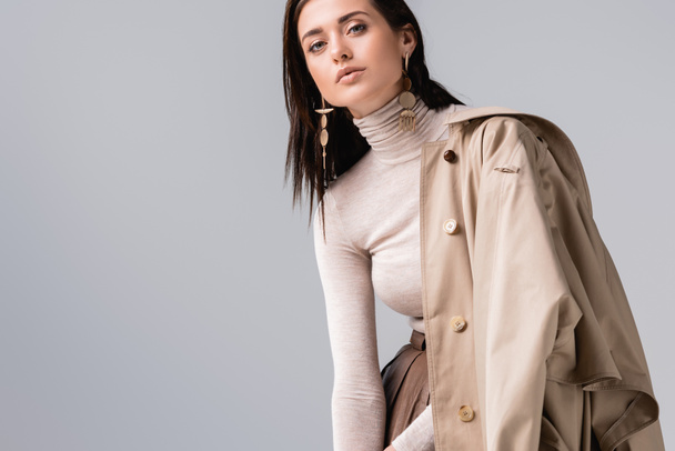 attractive, stylish girl touching collar of trench coat and looking at camera isolated on grey - Photo, Image