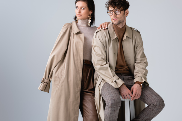 trendy woman looking away while touching shoulder of stylish man sitting on stool isolated on grey - Foto, imagen