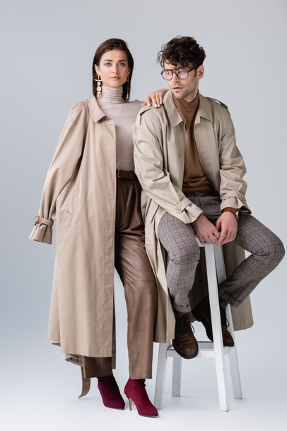 full length view of fashionable woman touching shoulder of trendy man sitting on grey - Foto, immagini