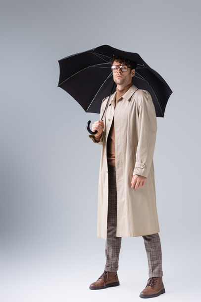 vue complète de l'homme confiant et élégant regardant la caméra tout en posant sous le parapluie sur le gris - Photo, image