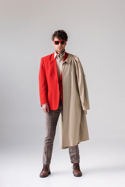 full length view of fashionable, confident man with trench coat over shoulder looking at camera on grey - Photo, Image