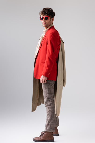 full length view of stylish man in red blazer holding trench coat and looking at camera on grey - 写真・画像