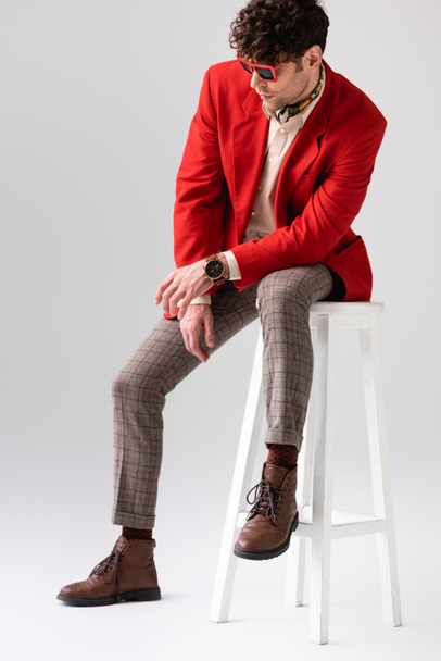 trendy man in red blazer and sunglasses sitting on stool with bowed head on grey - Zdjęcie, obraz