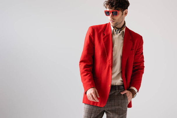 fashionable man in red blazer and sunglasses holding hand in pocket isolated on grey - Фото, изображение