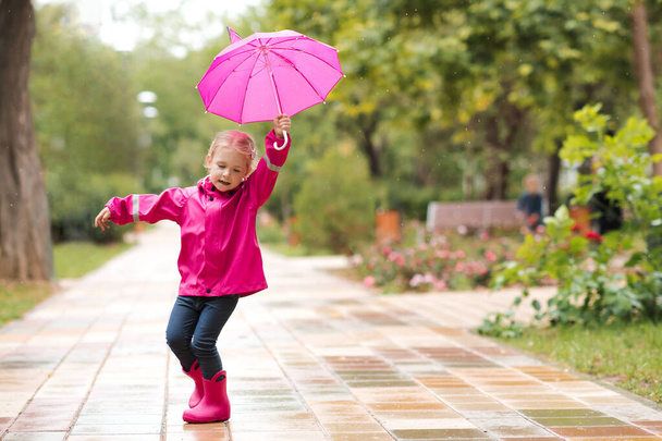 Девочка 2-3 лет ходит по луже в парке с розовым зонтиком в водонепроницаемых сапогах на открытом воздухе. Детство. Осенний сезон.  - Фото, изображение