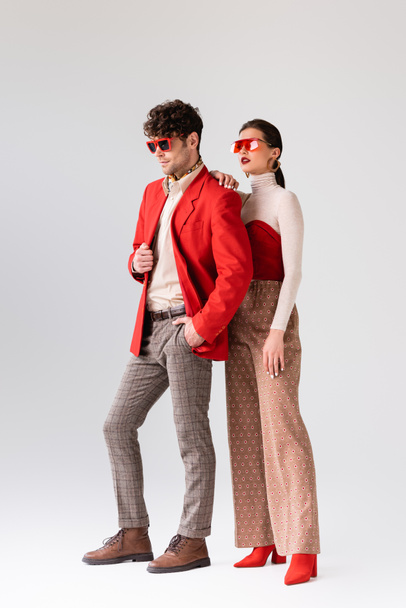 full length view of elegant girl touching shoulder of trendy man in red blazer on grey - Photo, Image