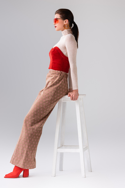 boční pohled na módní dívku v béžových kalhotách a červeném korzetu na roláku opírající se o stoličku na šedé - Fotografie, Obrázek