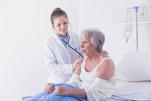 Γυναίκα γιατρός ακούει μια ηλικιωμένη γυναίκα ασθενείς πνεύμονες και την καρδιά με στηθοσκόπιο κατά τη διάρκεια μιας διαβούλευσης σε ένα θάλαμο νοσοκομείου, καθώς κάθεται στο κρεβάτι - Φωτογραφία, εικόνα