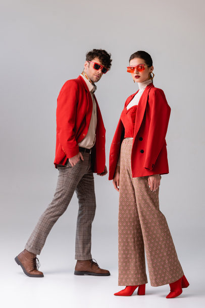 full length view of fashionable couple in red blazers and sunglasses looking at camera  on grey - 写真・画像