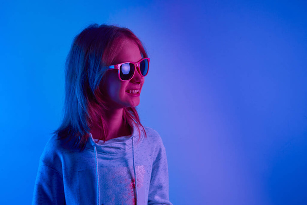 Ευτυχισμένο έφηβο κορίτσι στέκεται και χαμογελά πάνω από μοντέρνο μπλε νέον φως. Πορτρέτο μιας νέας, όμορφης έφηβης. Συνθετικό κύμα. - Φωτογραφία, εικόνα