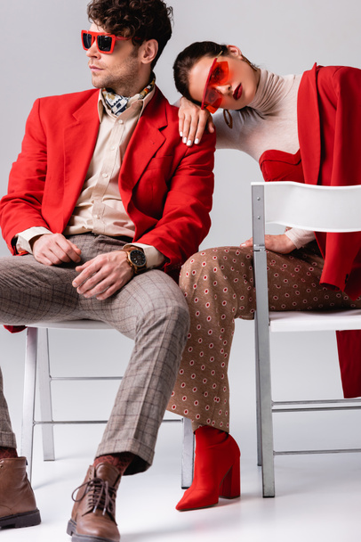 stylish woman leaning on shoulder of trendy man while sitting on chairs on grey - Φωτογραφία, εικόνα
