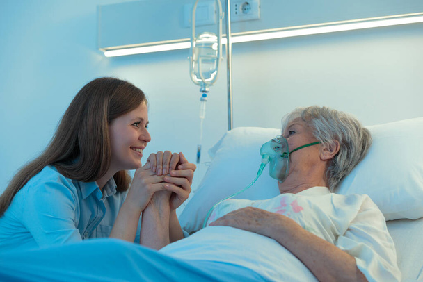 Young woman visiting her elderly mother or grandmother in hospital as she lies in a bed using a positive pressure oxygen mask to aid her breathing with a comforting positive smile as she clasps her hand - Photo, Image