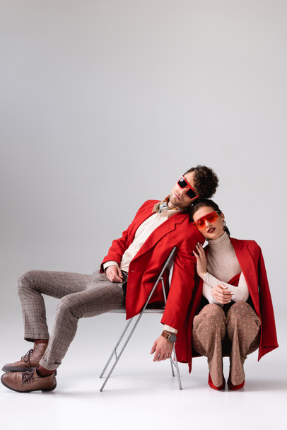 elegant woman squatting and touching trendy man sitting on chair on grey - Photo, Image