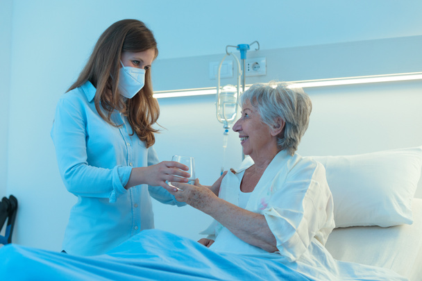 Cuidador o hija pequeña que lleva una mascarilla que le da a una anciana sonriente medicación para pacientes y un vaso de agua mientras se sienta en la cama de un hospital - Foto, Imagen