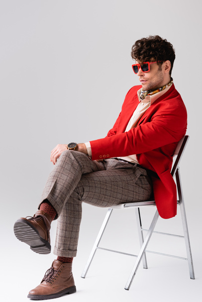 Stylish man in red blazer with crossed legs, sitting on chair on grey - Fotó, kép