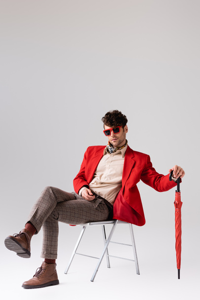 Stylish man in red blazer with crossed legs, posing with umbrella while sitting on chair on grey - Fotó, kép